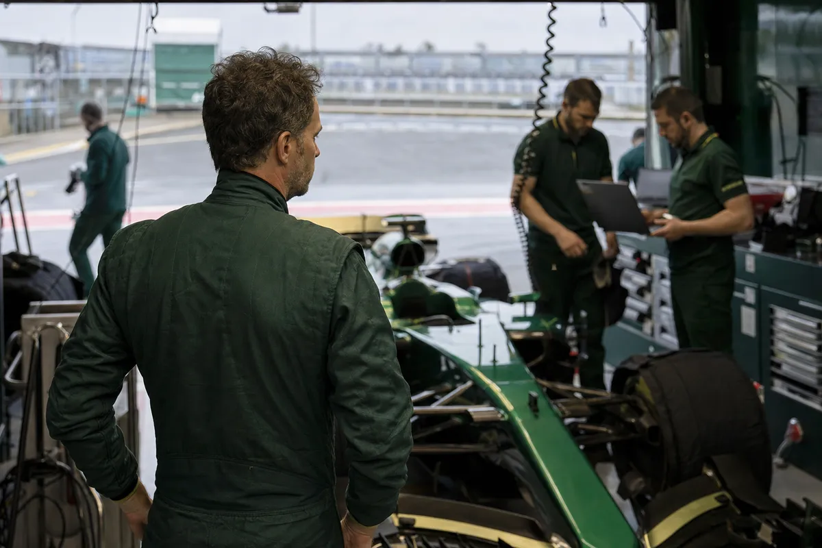 Pilota in tuta verde osserva una monoposto verde nel box, con ingegneri al lavoro con laptop