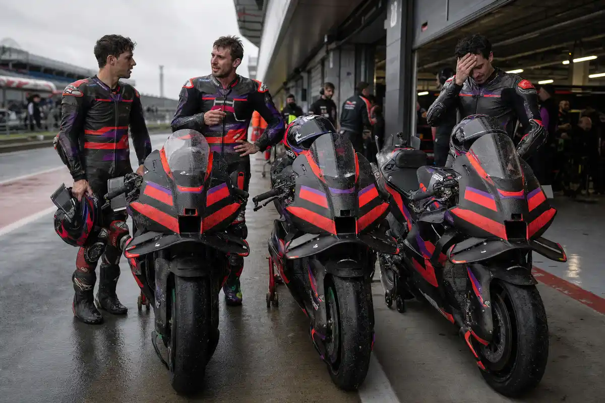 Tre moto da gara rosso-nero-viola in pit lane bagnata, con tre piloti in tuta accanto al box dopo la gara