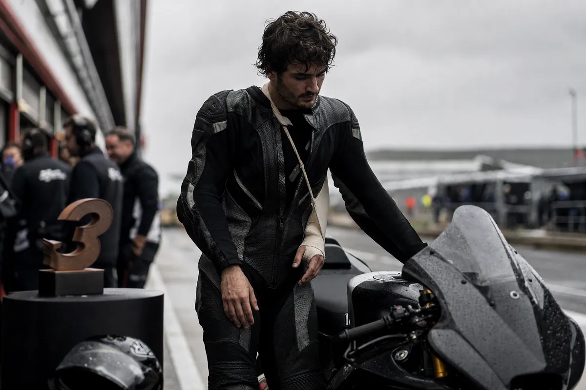 Pilota di MotoGP stanco in tuta nera, con braccio fasciato, accanto alla moto nel box su pista bagnata