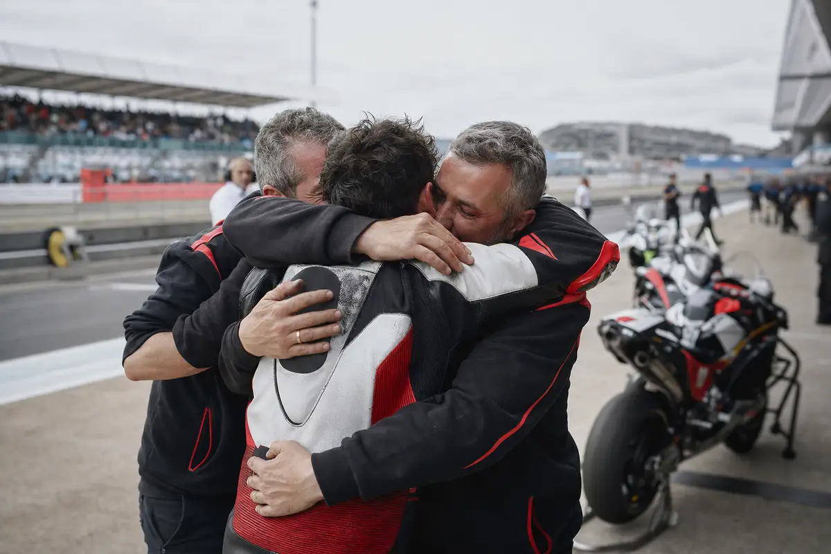 Pilota di motociclismo abbracciato da due membri del team nel paddock, con moto da gara sullo sfondo