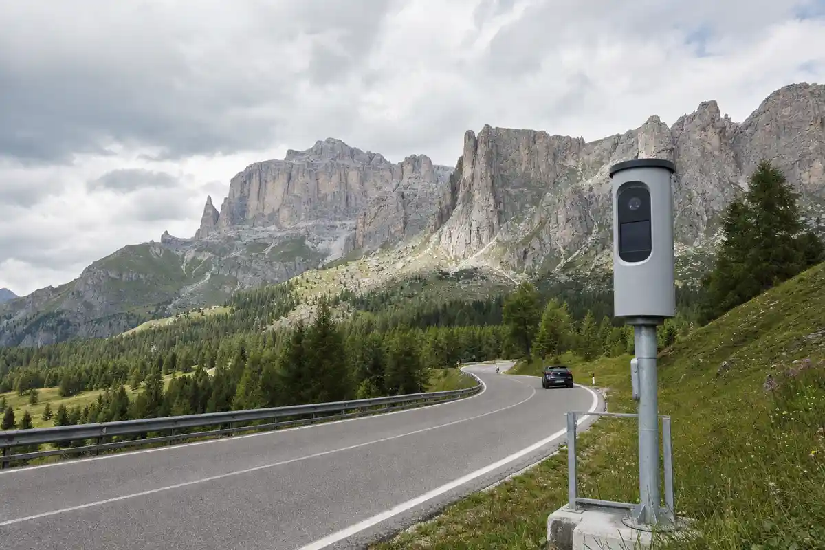 Autovelox a bordo strada su curva di montagna con auto in lontananza e cime dolomitiche sullo sfondo
