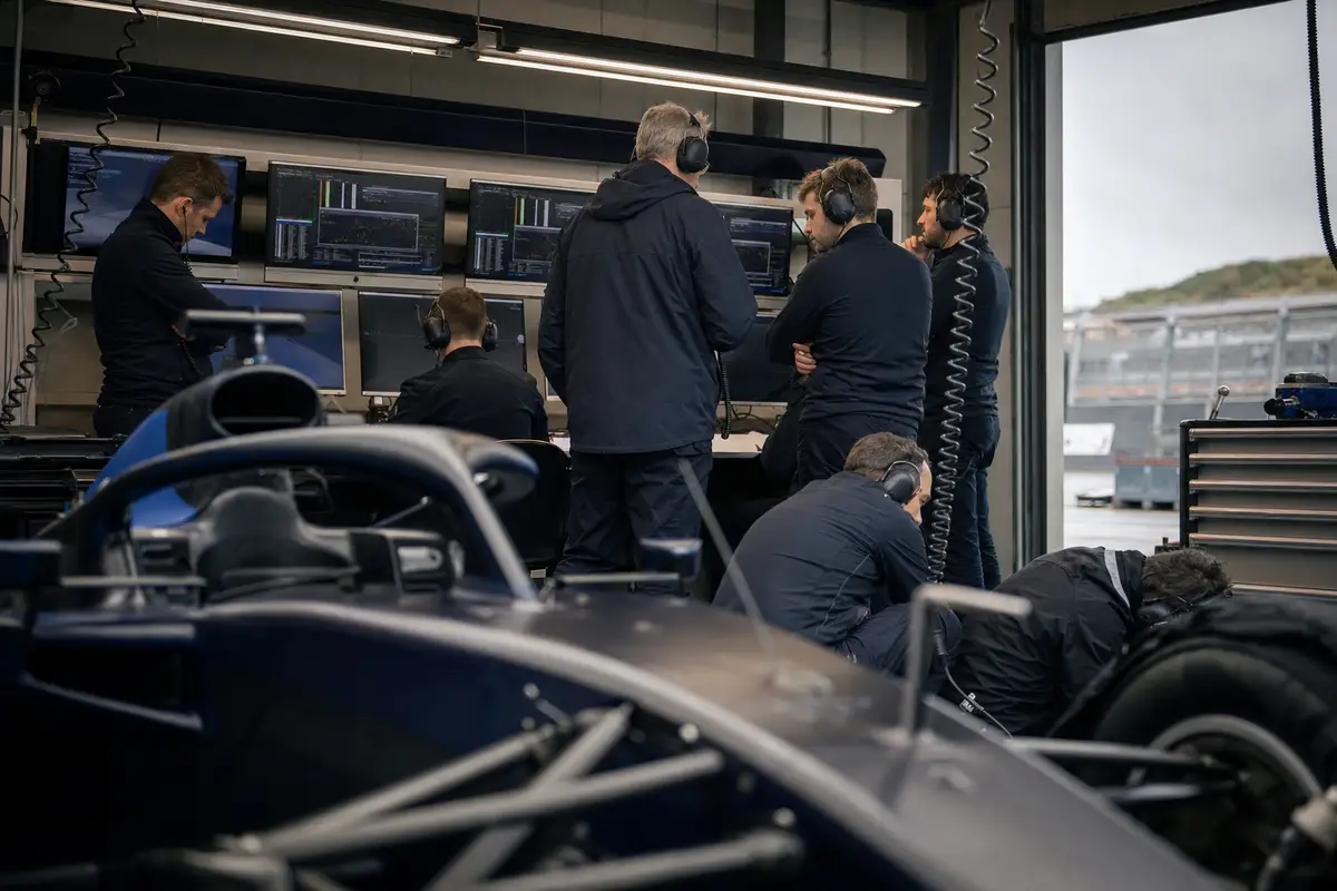 Ingegneri in un box di Formula 1 davanti ai monitor, con una monoposto scura sfocata in primo piano
