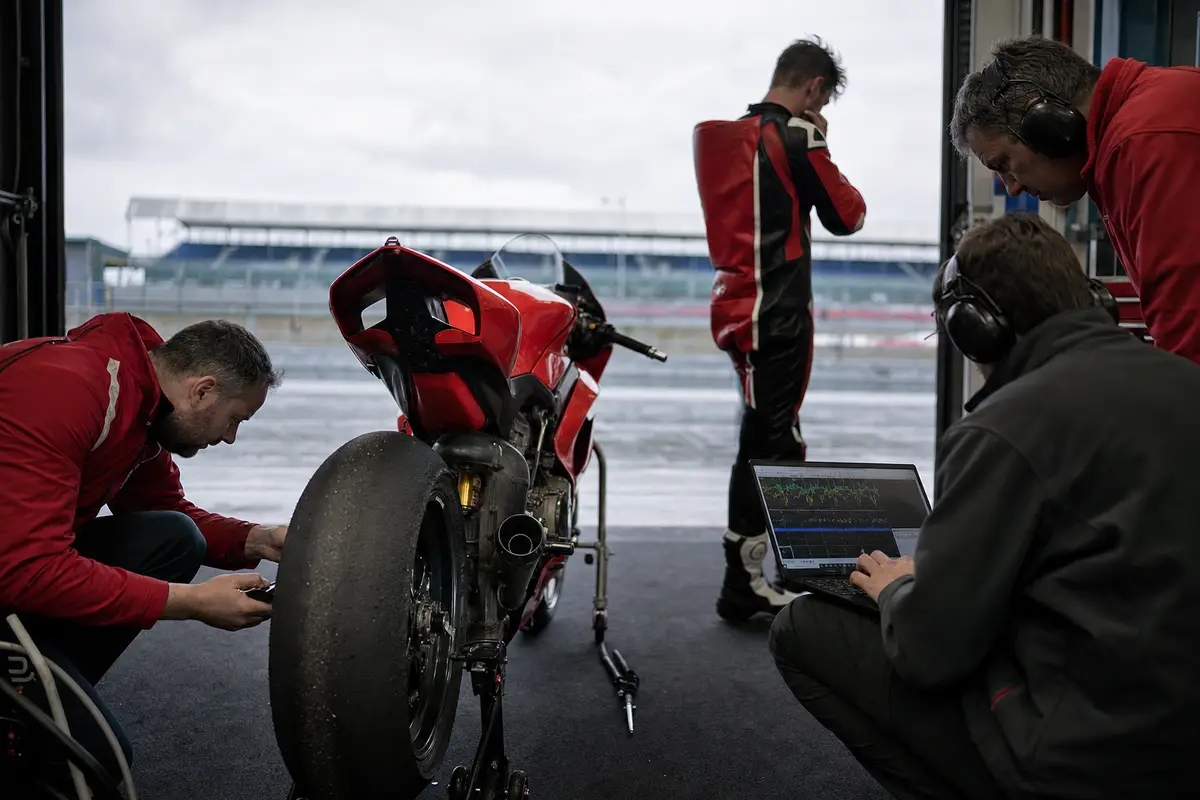Moto da gara rossa ai box, tecnico controlla la gomma posteriore mentre un altro analizza dati al laptop