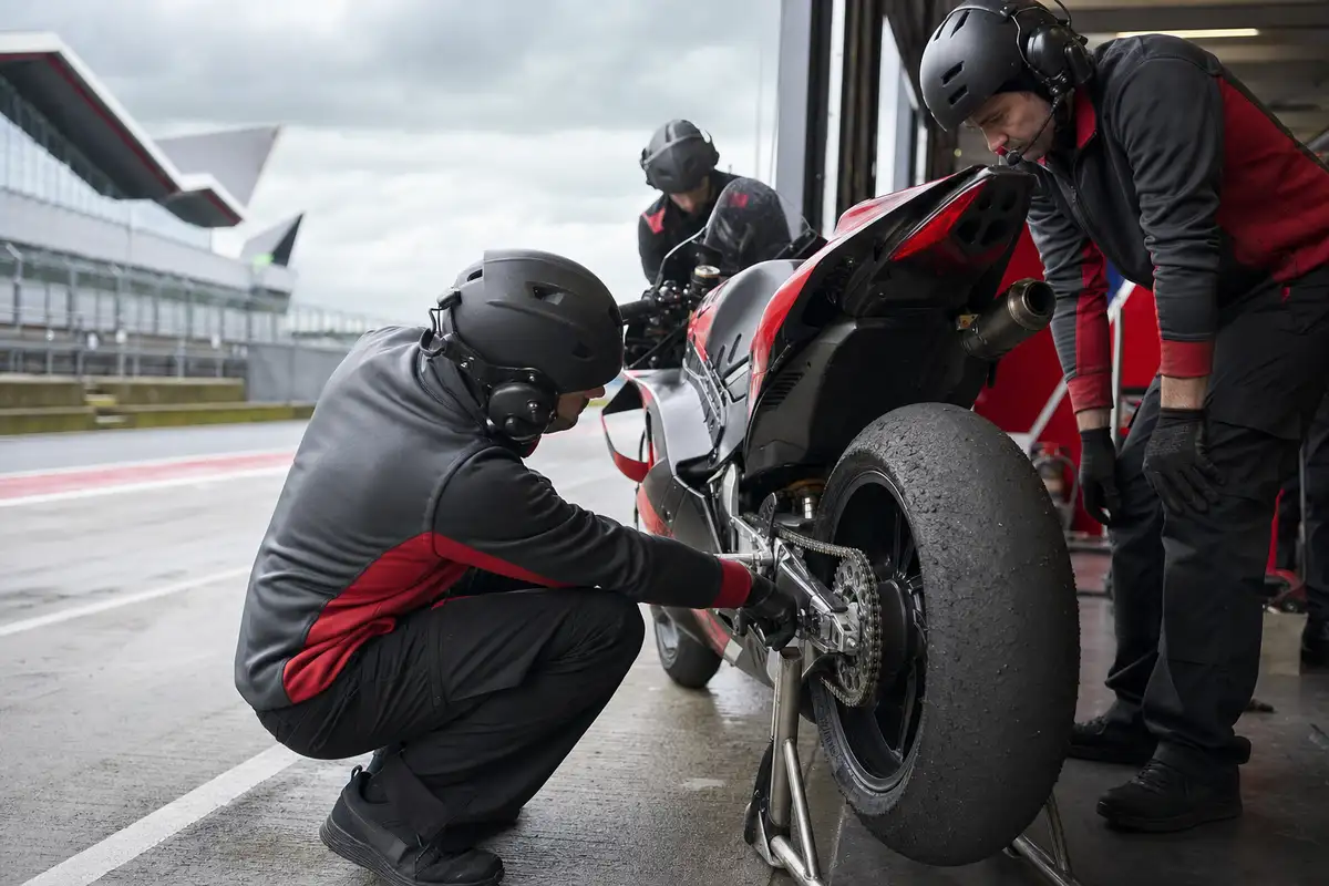 Meccanici controllano la gomma posteriore consumata di una moto da gara nel box ai box, con pista bagnata sullo sfondo