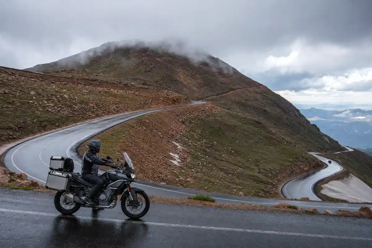 Motociclista su enduro stradale con borse laterali, fermo su asfalto bagnato lungo una strada di montagna tra nuvole