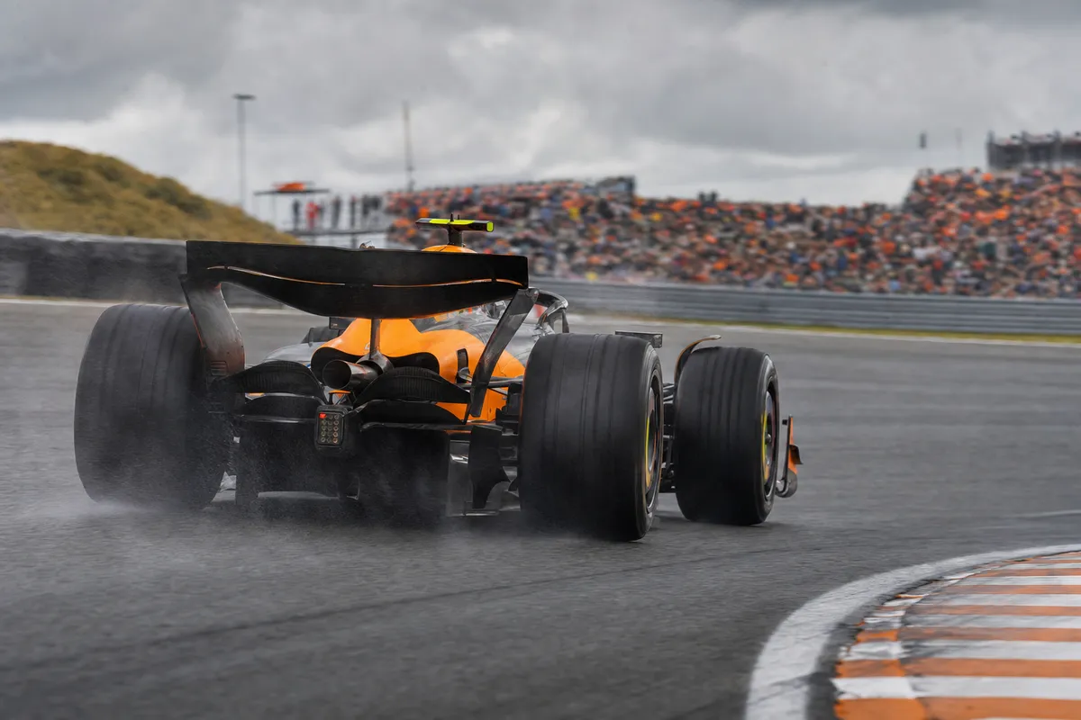 Monoposto arancione e nera in uscita di curva su pista bagnata, con spray dagli pneumatici e tribune sfocate