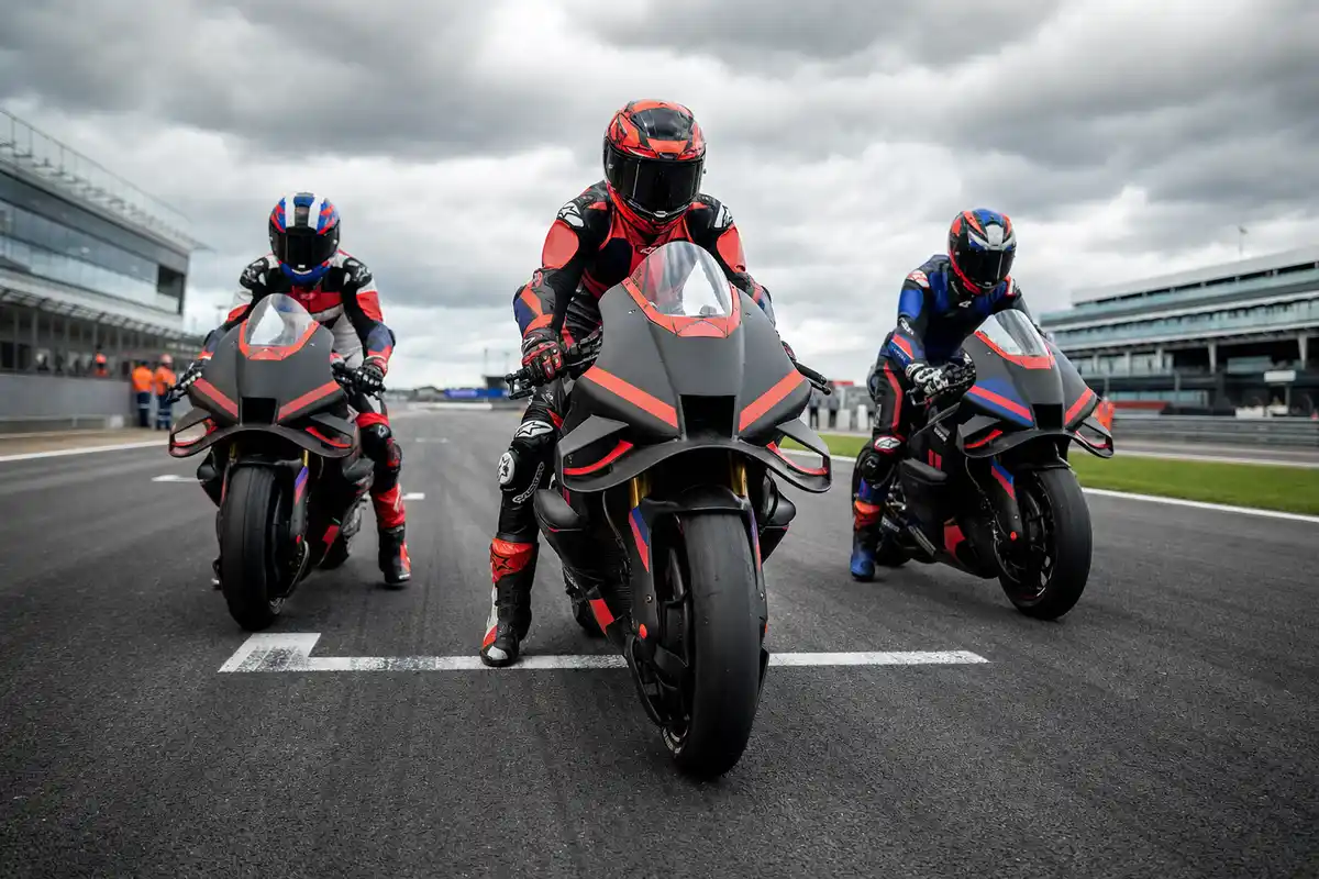 Tre moto da gara con piloti in tuta e casco schierati in prima fila sulla griglia, cielo nuvoloso