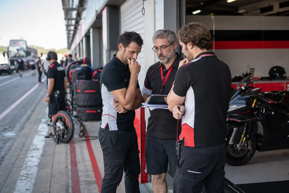 Tre membri del team discutono guardando un tablet davanti a un box in pit lane, con una moto da gara sullo sfondo