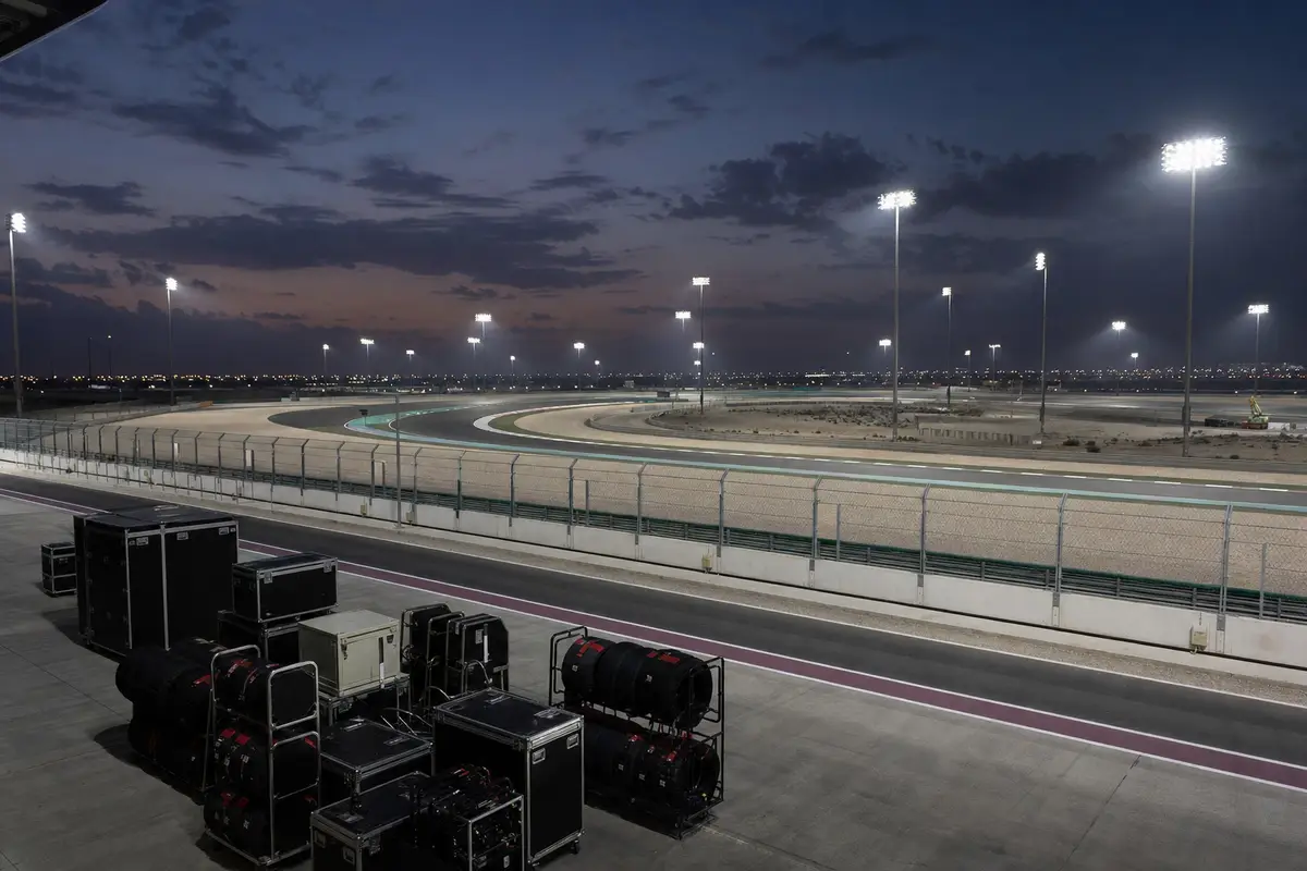 Pit lane vuota con casse e gomme accatastate davanti a un circuito nel deserto illuminato dai riflettori al crepuscolo