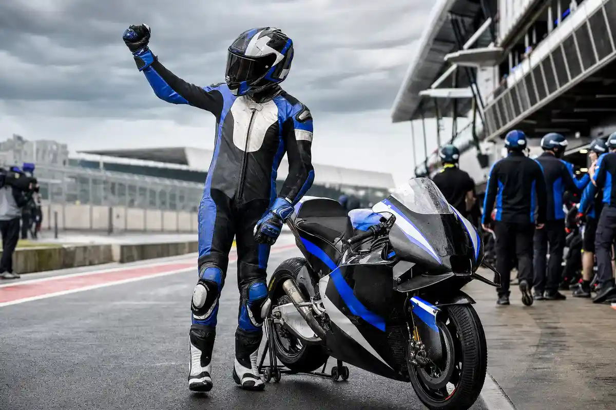Pilota in tuta blu e nera alza il pugno accanto a una moto da corsa nel paddock, cielo coperto sullo sfondo