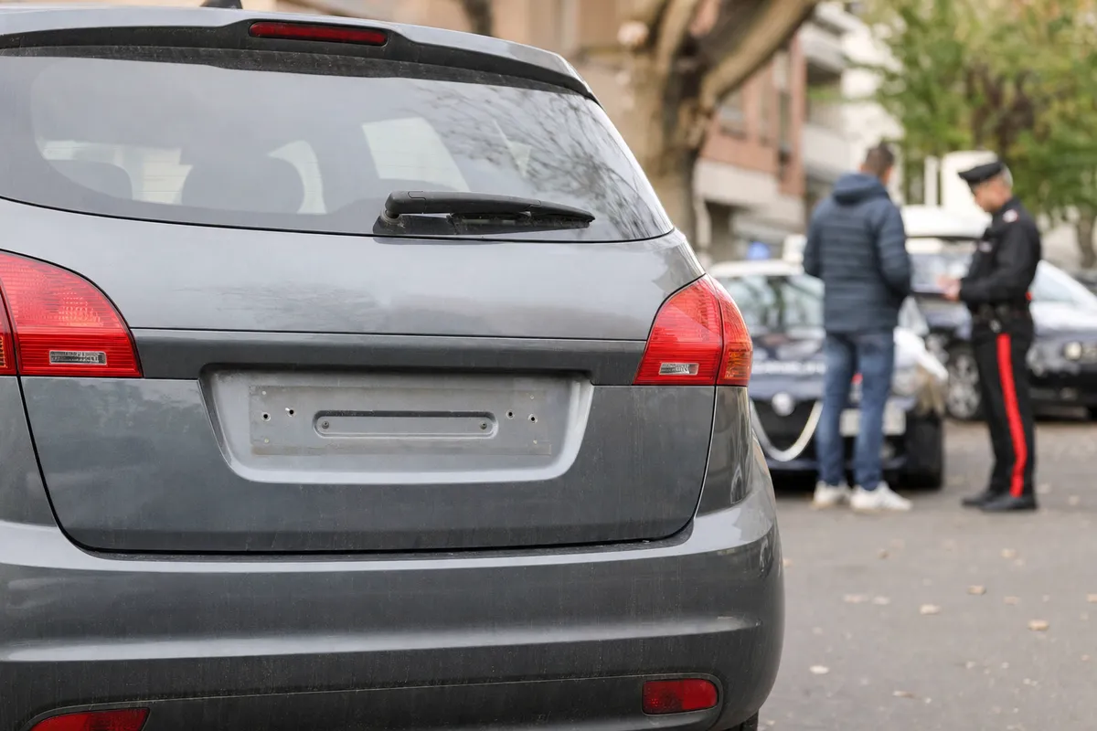 Retro di un’auto grigia parcheggiata con portatarga posteriore vuoto; sullo sfondo un agente parla con un uomo