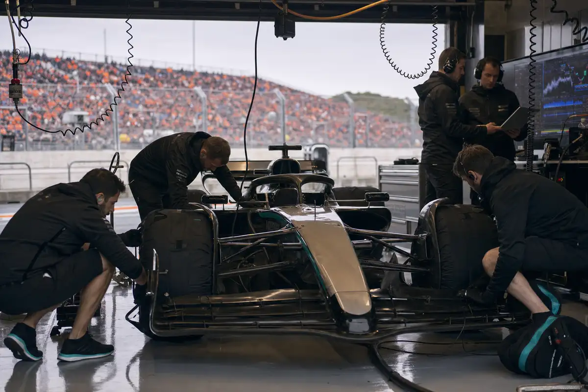 Meccanici al lavoro su una monoposto di Formula 1 argento e nera nel box, con tribune arancioni sullo sfondo