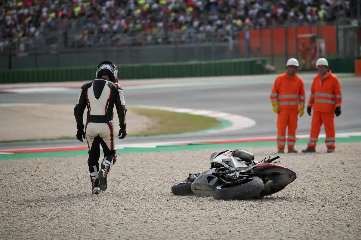 Moto da corsa a terra nella ghiaia e pilota che si allontana, con due commissari in tuta arancione