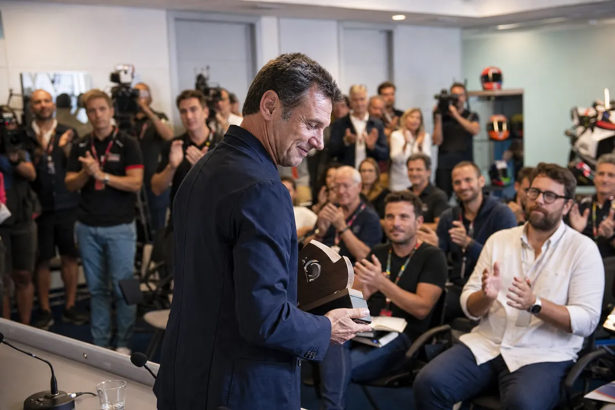 Uomo con targa commemorativa in sala stampa, mentre giornalisti e addetti ai lavori applaudono