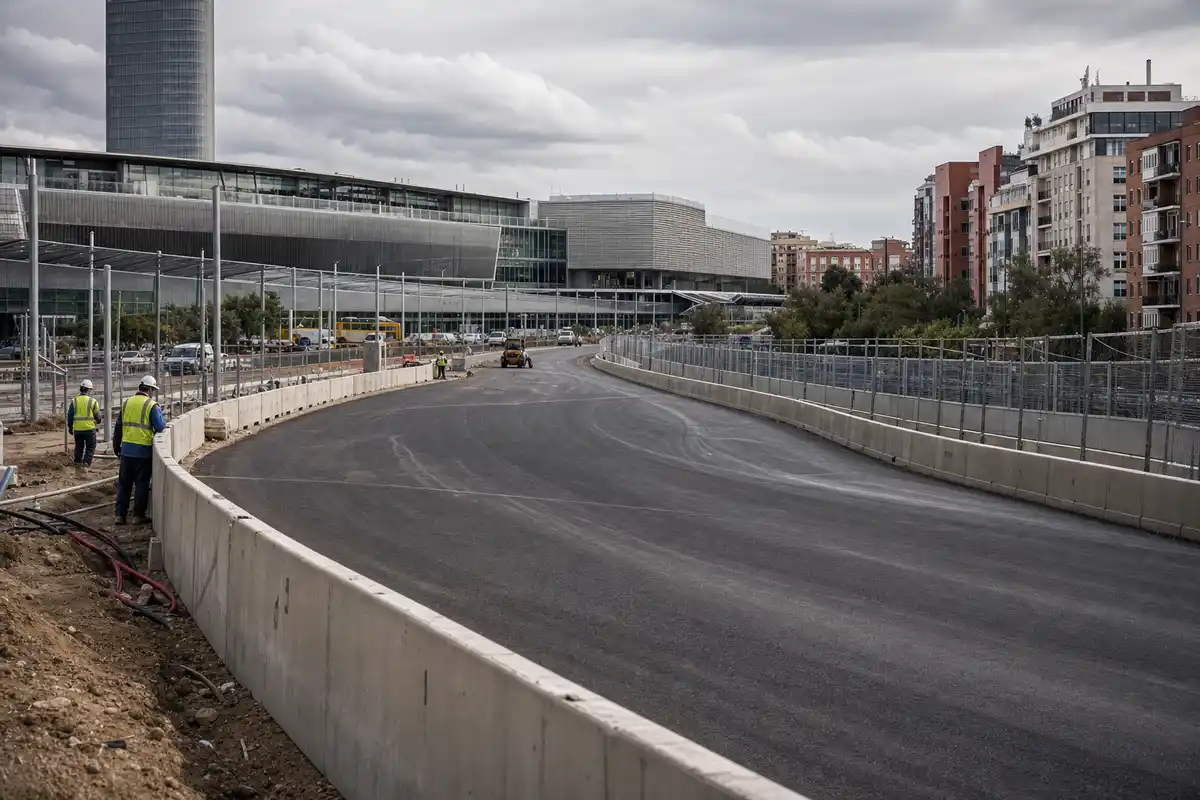 Curva di un circuito cittadino in costruzione con barriere in cemento, reti metalliche e operai davanti a palazzi moderni