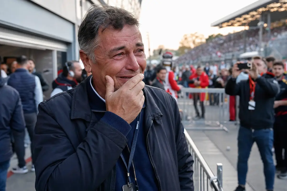 Uomo commosso nel paddock di un circuito, mano alla bocca, con folla e tribune sullo sfondo