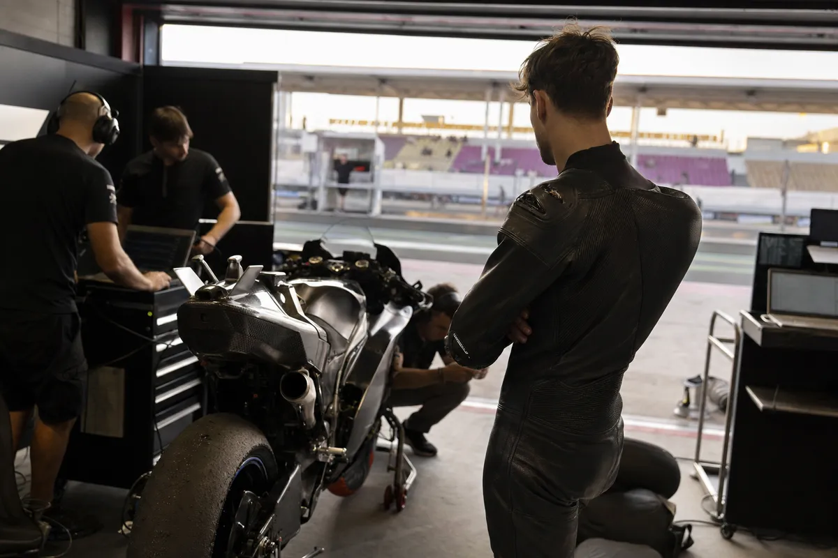 Pilota in tuta nera a braccia conserte accanto a una moto da gara nel box, con meccanici e laptop sullo sfondo
