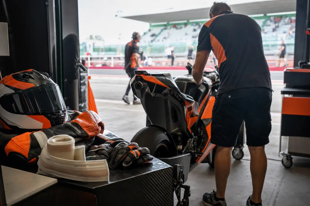 Tuta e casco da MotoGP su una panca con fascia elastica e guanti, mentre un meccanico lavora su una moto in box