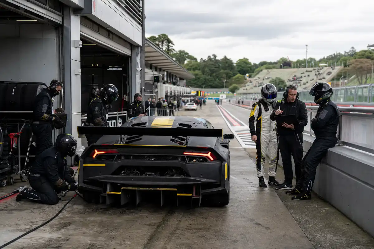 Auto GT da endurance ai box con meccanici e piloti, sullo sfondo pit lane e tribuna